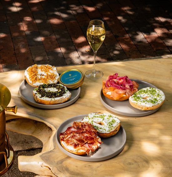 A rustic wooden table holds plates of bagels topped with varied ingredients like caviar, salmon, and ham, paired with a glass of champagne, under dappled sunlight.