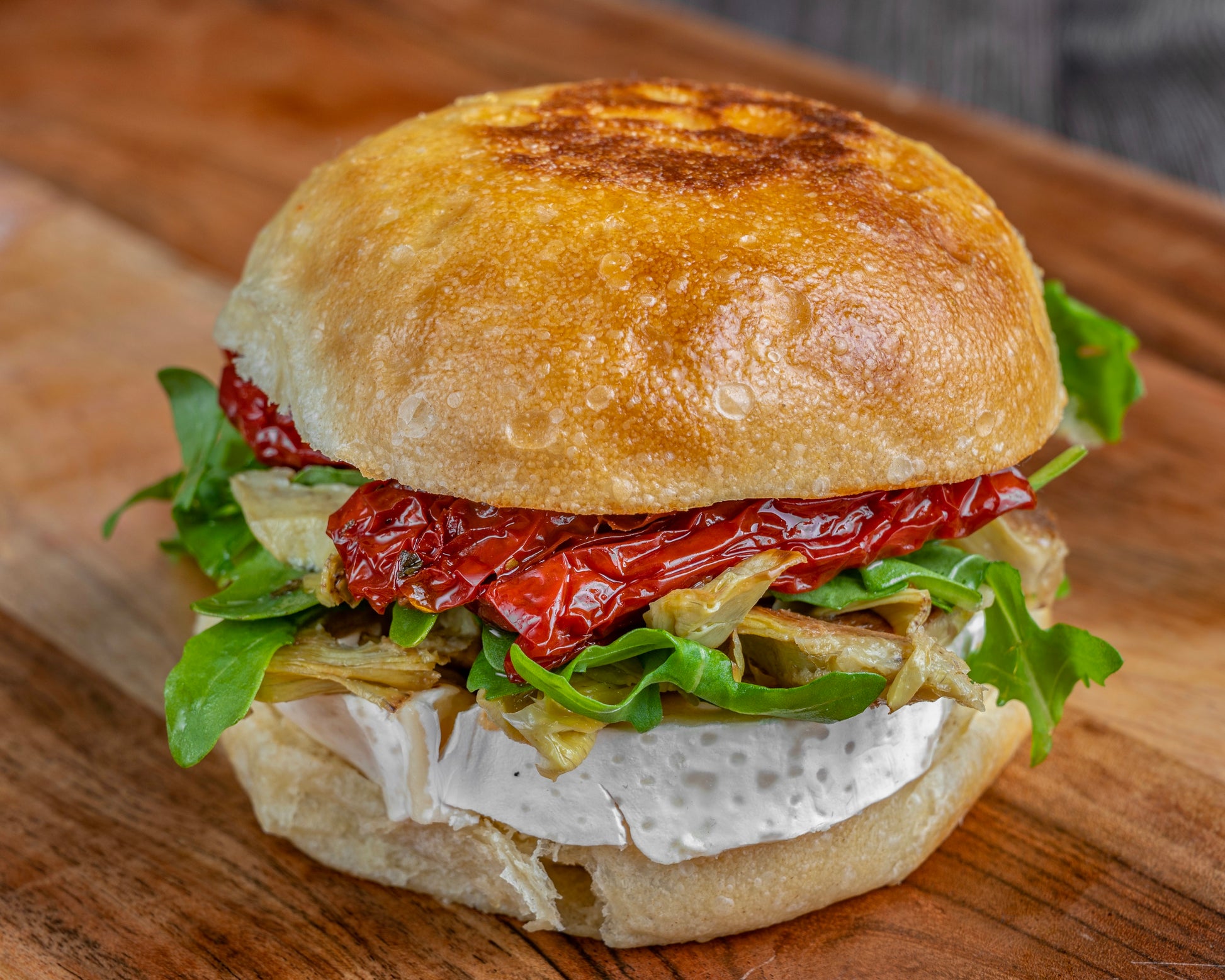 A gourmet sandwich on a wooden board, featuring a crusty bun, creamy Brie, arugula, sun-dried tomatoes, and roasted artichokes, creating a fresh, savory look.